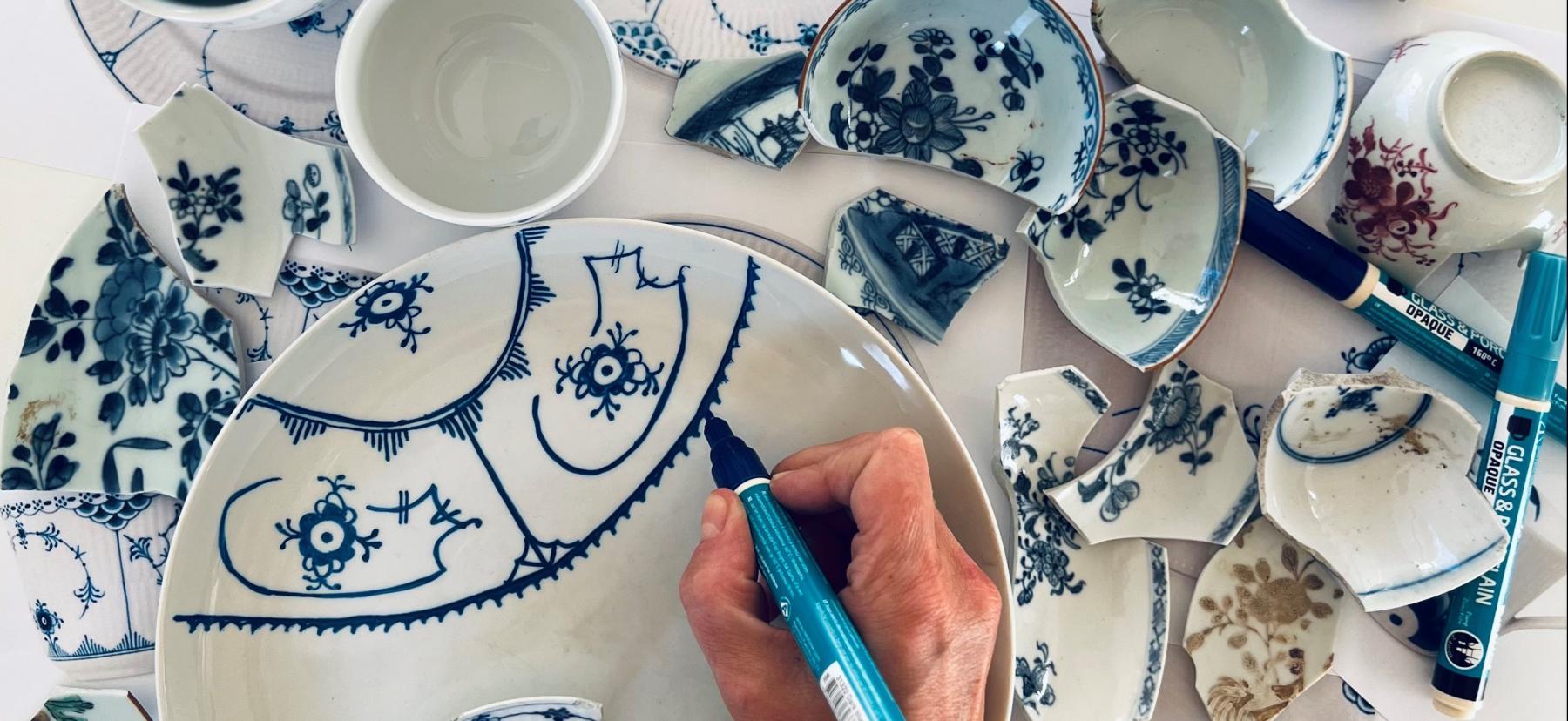 Hånd, der dekorerer hvidt porcelæn med blå tusch-maling.