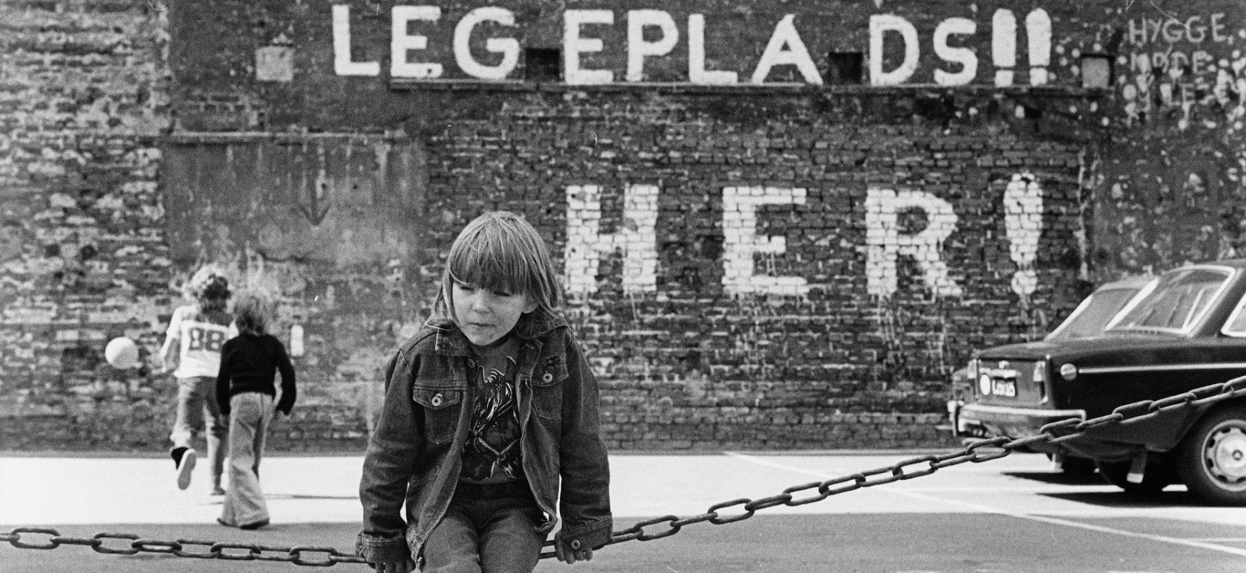 Sort/hvid foto af barn, foran et murmaleri med teksten Legeplads Her!