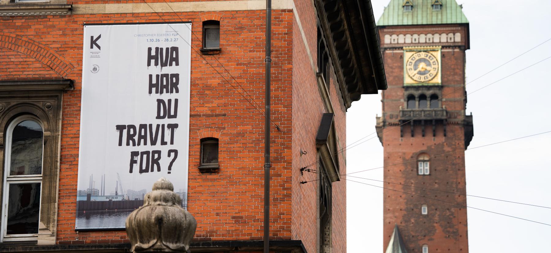 Udstillingsplakat med teksten HVA' HAR DU TRAVLT FOR? på Københavns Museums facade. Rådhustårnet i baggrunden